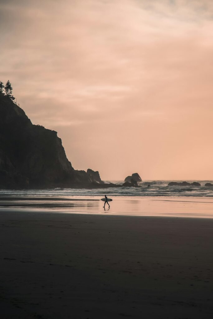 A lone surfer walks along the serene Oregon beach at sunset, carrying a surfboard.