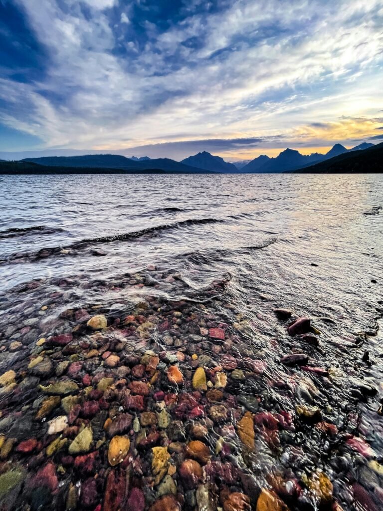 A tranquil view of Lake McDonald with colorful rocks and distant mountains at dusk.