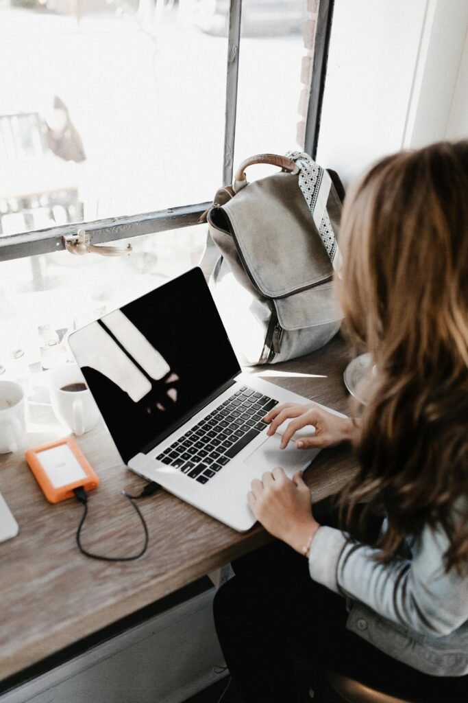 A young woman works remotely at a café, using her laptop and external hard drive.