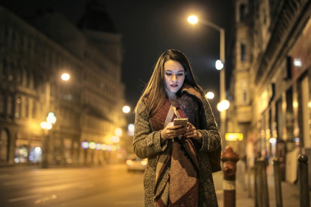 Elegant woman walks through urban street at night, engaged with her smartphone, surrounded by city lights.