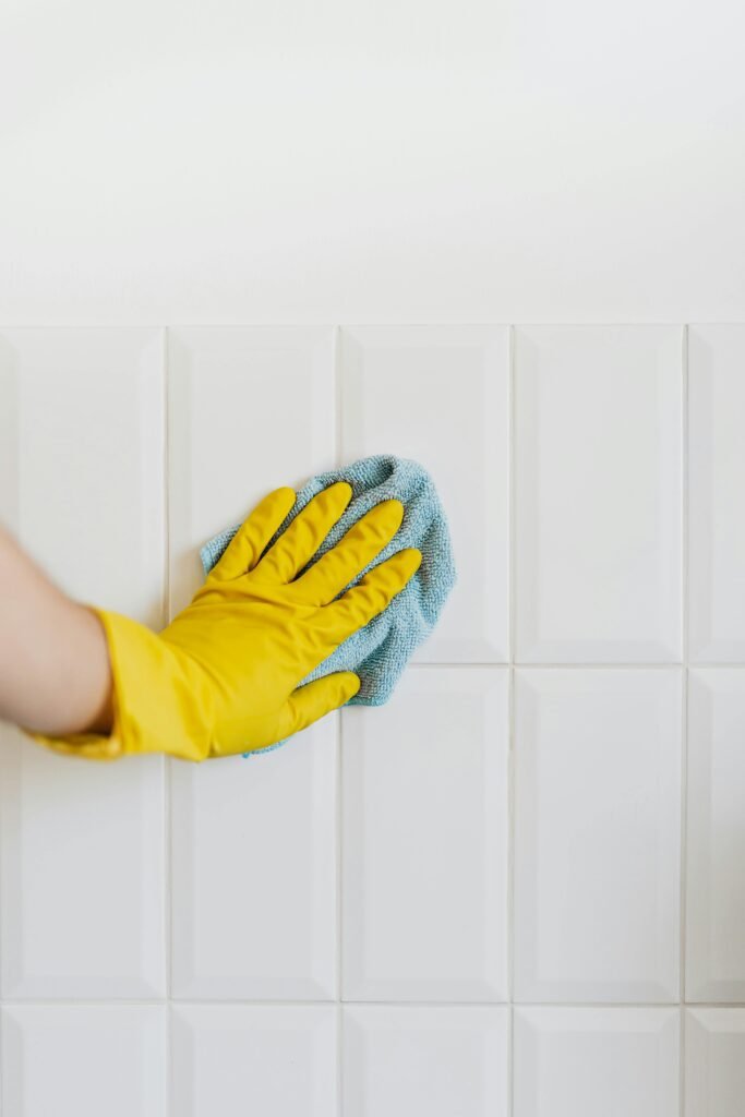 Close-up of a gloved hand wiping a white tiled wall with a blue cloth, symbolizing cleanliness and hygiene.