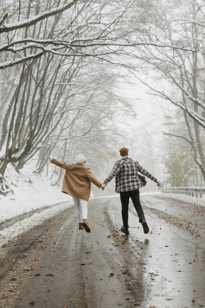 A couple joyfully walking hand in hand on a snowy winter road, surrounded by trees.