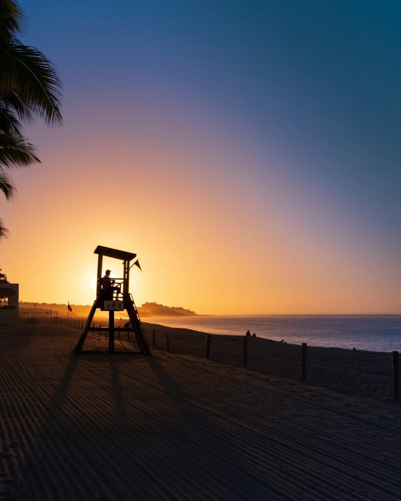 Beautiful silhouette of a lifeguard tower at Cabo San Lucas beach during sunrise, showcasing ocean and palm trees.