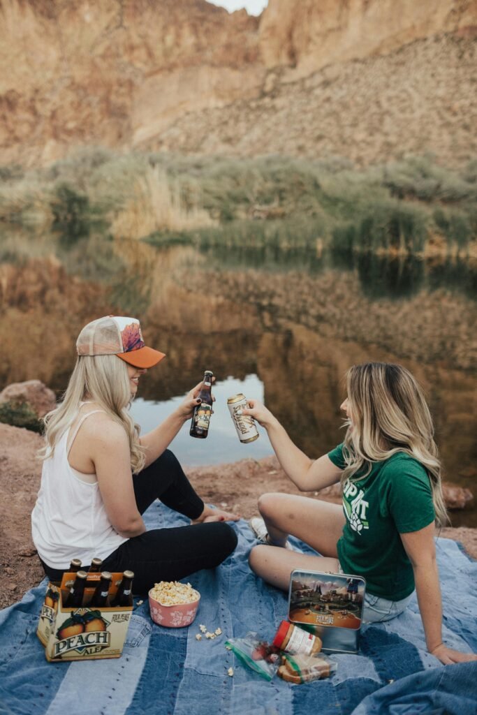 Two women having a joyful picnic by a picturesque lakeside, drinking and enjoying snacks.