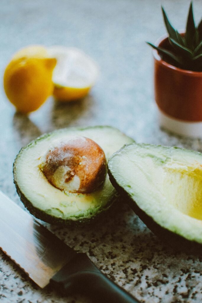 Close-up of a sliced avocado with knife, lemon and potted plant on a kitchen counter.