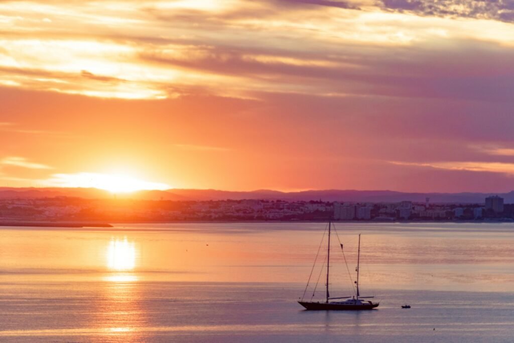 A serene sunset view over Lagos, Portugal with a silhouetted sailboat on the calm sea.