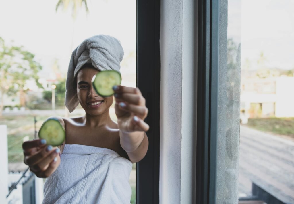 Smiling woman with towel turban holding cucumber slices, enjoying a skincare routine by the window.